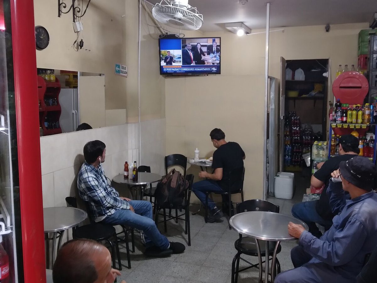 Inside a small neighbourhood shop and bar in Medellín: customers seated at metal tables watch a television, with a drinks fridge and shelves of bottled drinks
